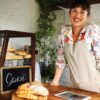 Mulher sorridente com uniforme atrás de balcão de uma padaria local, simbolizando como empreender com pouco dinheiro